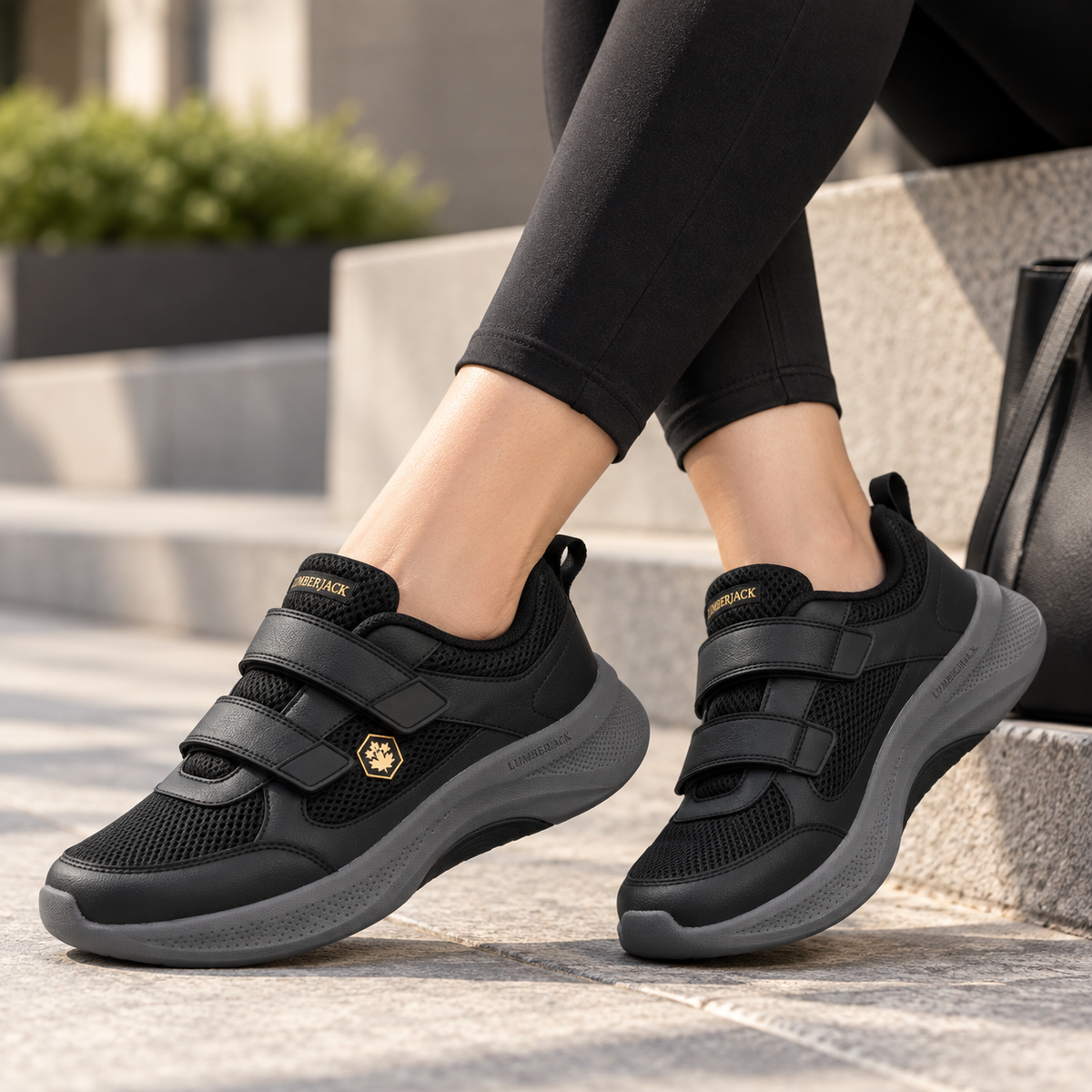 A person wearing black low-top sneakers with hook-and-loop straps and a small gold logo, photographed sitting on outdoor stone steps.