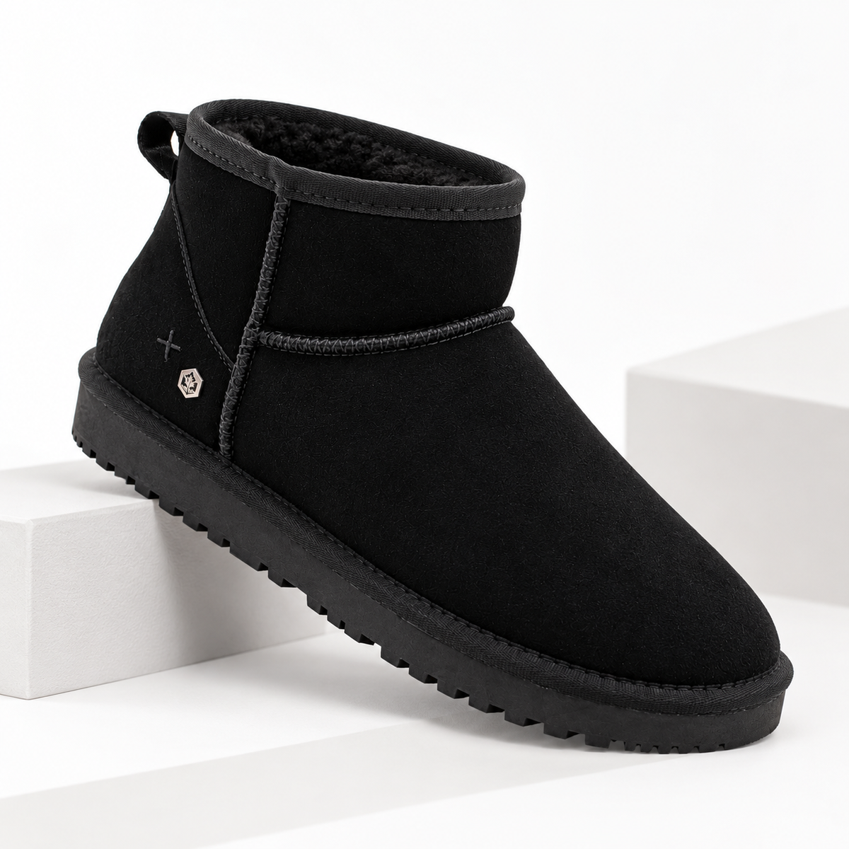 A single black suede ankle boot with a thick black sole, shown resting on a white geometric platform against a white background.
